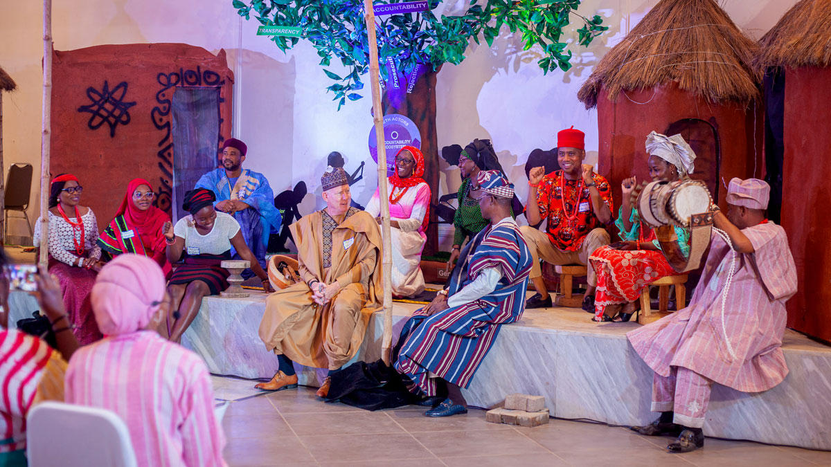 A group of people in traditional attire seated on a platform in a village-like setting with huts and decorative elements.